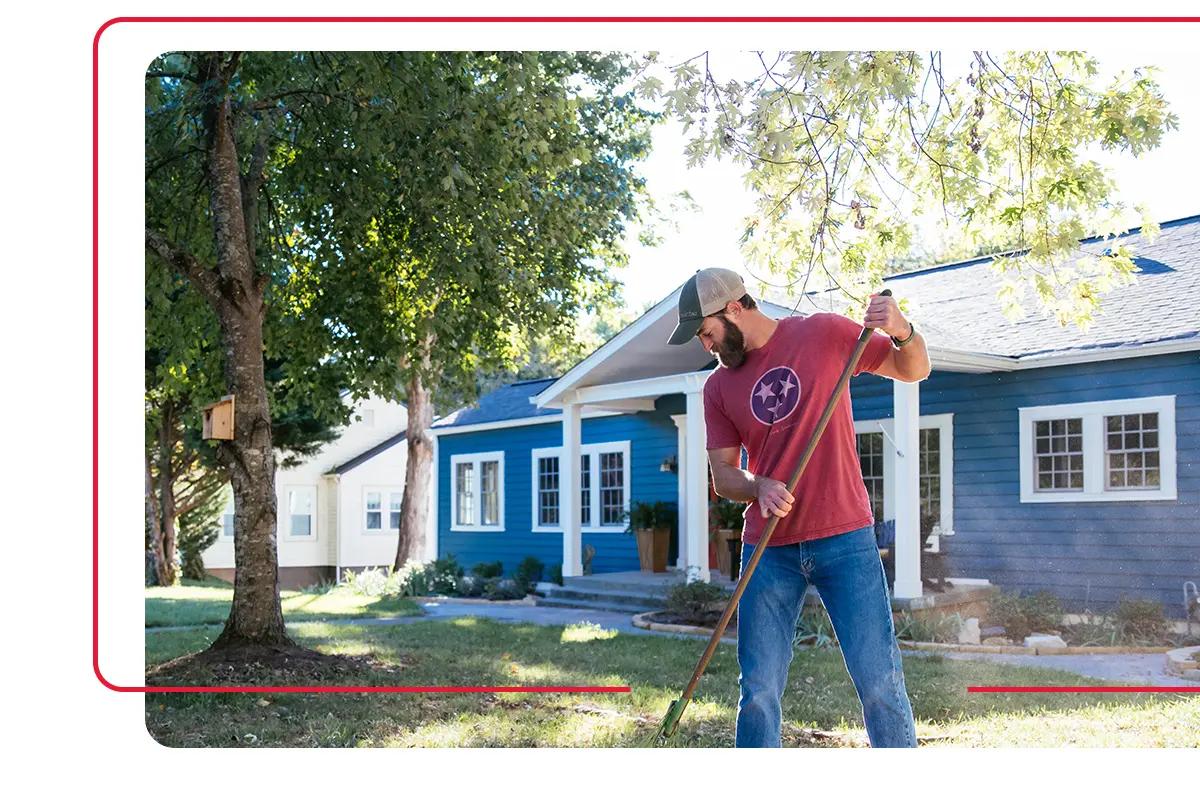 Homeowner in red Tennessee tri star logo shirt using rake to care for the front lawn of the blue home he owns