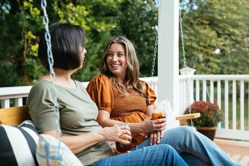 Two women sitting on front porch swing holding mason jars of iced tea