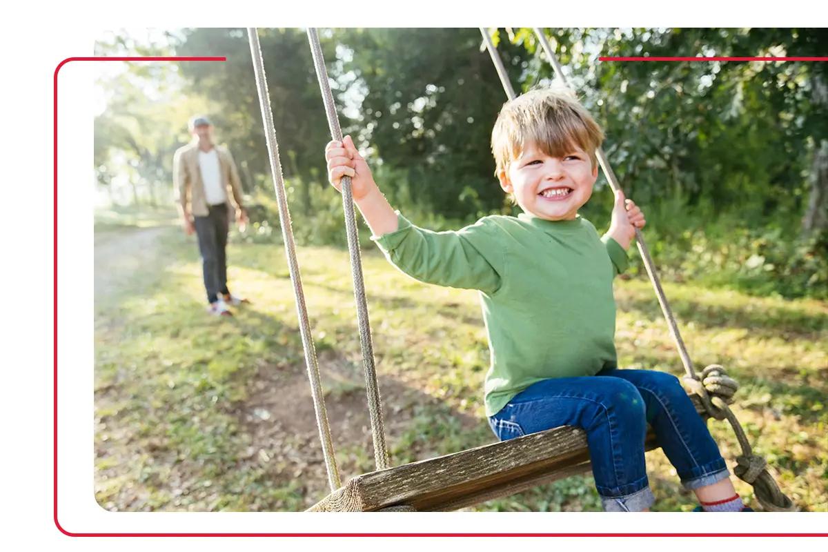 Young child in green shirt on tree swing with his father pushing him in the background