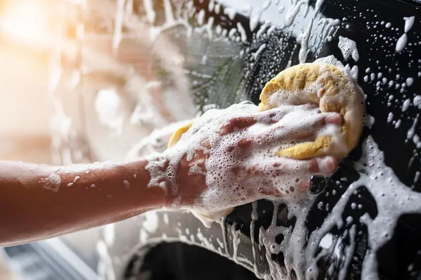 Car owner hand-washing a black car in the driveway with a sponge and soap